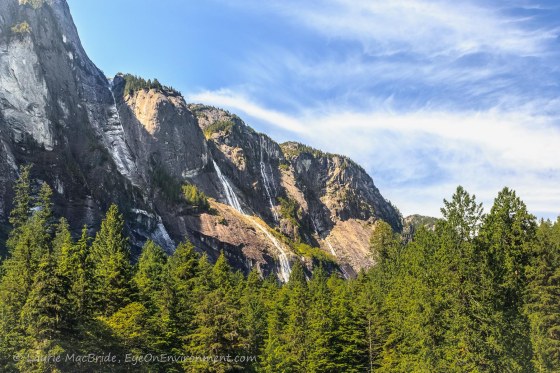 Light and shadow over high elevation mountains, with waterfalls and forest below.